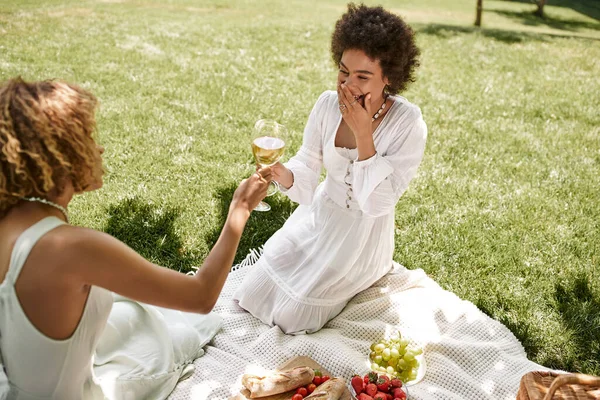 Mujer afroamericana riendo y tintineando copas de vino con novia cerca de aperitivos, picnic de verano - foto de stock