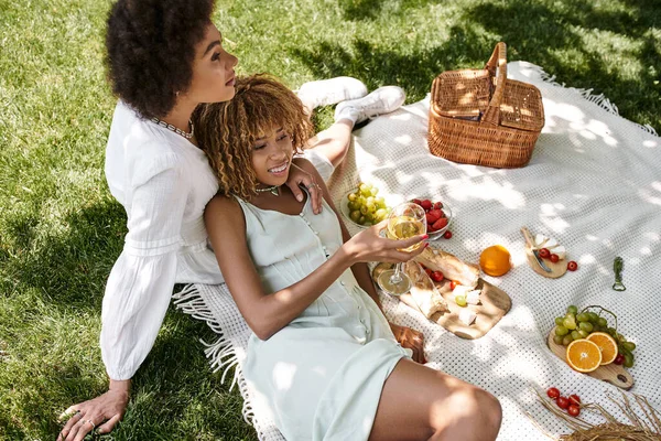 Sonriente mujer afroamericana sosteniendo copa de vino cerca de su novia, relajación en el picnic de verano - foto de stock