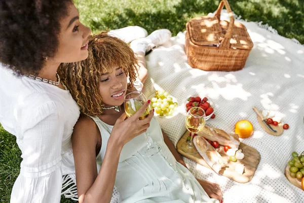 African american woman drinking wine near girlfriend and snacks on blanket, summer picnic — Stock Photo