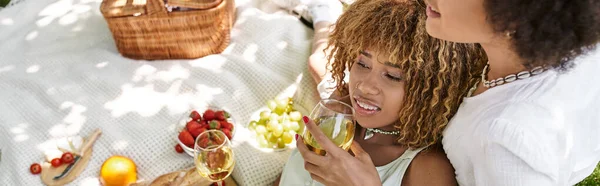 Felice donna africana americana con bicchiere di vino vicino fidanzata e cibo sulla coperta, tempo libero estivo — Foto stock