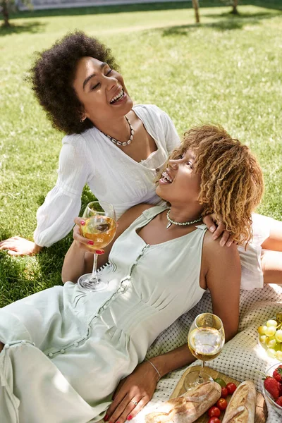 Беззаботная африканская американка смеется возле подружки с бокалом вина на пикнике в парке — стоковое фото