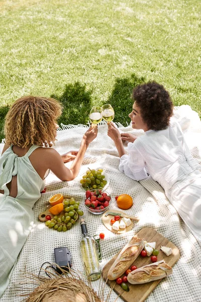 Novias afroamericanas tintineo copas de vino cerca de aperitivos en manta, picnic de verano - foto de stock