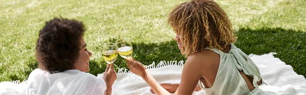 Happy african american girlfriends clinking wine glasses on blanket and green lawn, banner — Stock Photo