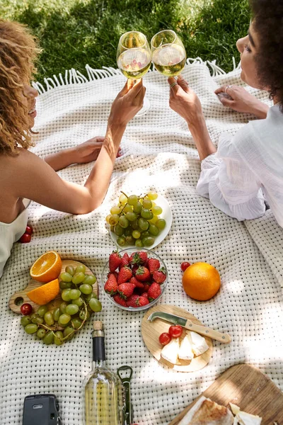 Fresh fruits and vegetables near african american girlfriends clinking wine glasses on picnic — Stock Photo