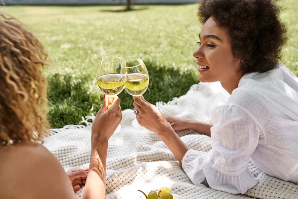 Alegre africana americana mujer tintineo copas de vino con novia en manta durante el picnic - foto de stock