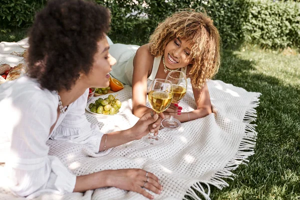 Sonriente afroamericana mujer tintineo copas de vino con novia en manta, ocio en el parque - foto de stock