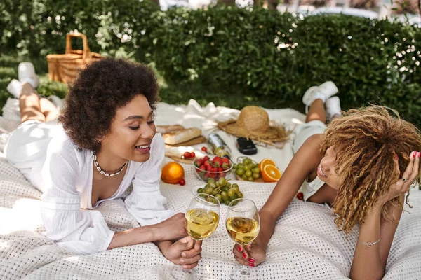African american girlfriends with wine glasses laying near fresh fruits and vegetables on blanket — Stock Photo