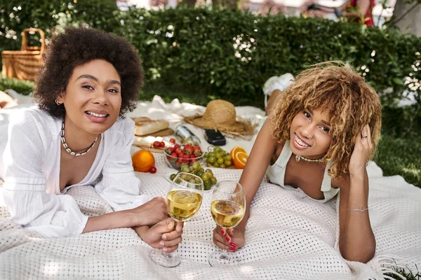 Молоді афроамериканські жінки з винними окулярами посміхаються на камеру біля свіжих закусок, літній пікнік — стокове фото