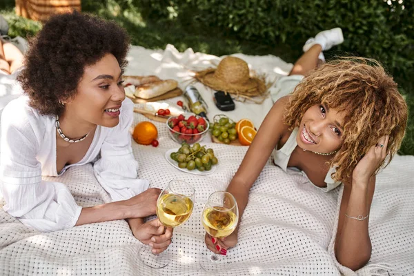 Mujer afroamericana con copa de vino mirando a la cámara cerca de la novia y aperitivos en el picnic de verano - foto de stock