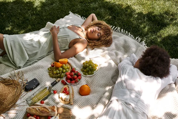 Smiling african american woman laying with closed eyes near girlfriend and snacks on blanket — Stock Photo