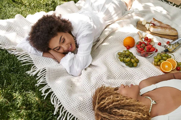 Fröhliche afrikanisch-amerikanische Frau liegt neben Freundin und frischem Obst beim Picknick im Park — Stockfoto