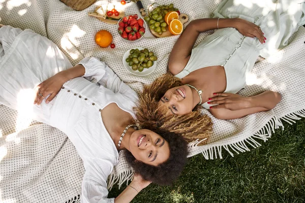 Heureuses copines afro-américaines allongées près de collations sur la couverture et regardant la caméra, vue de dessus — Photo de stock