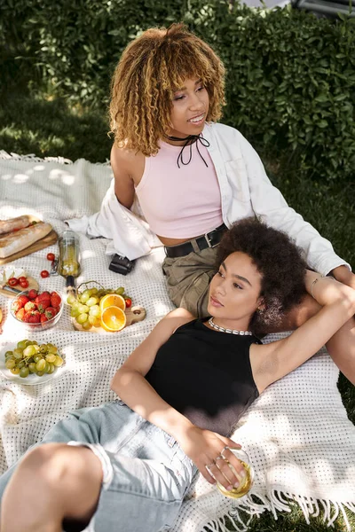 Mujer afroamericana sonriendo cerca de su novia en manta, picnic de verano, bocadillos, vino, relajarse - foto de stock