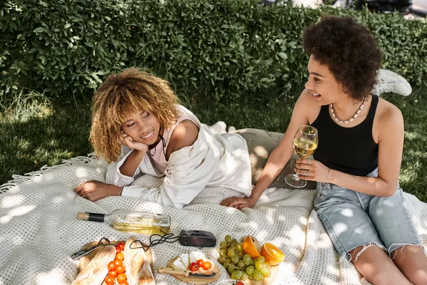 Молодая африканская американка с бокалом вина рядом с улыбающейся подругой и закусками, летний отдых — стоковое фото