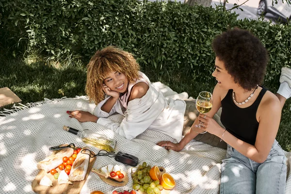 Mujer afroamericana con copa de vino hablando con su novia cerca de frutas y verduras en el picnic - foto de stock