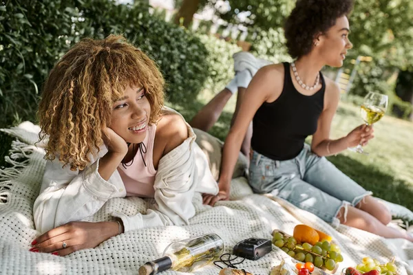 Sorridente mulher afro-americana olhando para longe perto de vinho, lanches e namorada no parque de verão — Fotografia de Stock