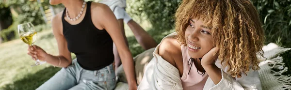 Sonriente mujer afroamericana mirando hacia otro lado cerca de novia con copa de vino en el picnic de verano, pancarta - foto de stock