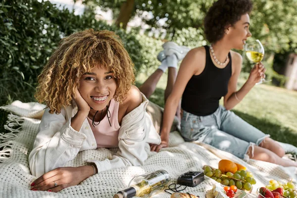 Pique-nique d'été, joyeuse femme afro-américaine regardant la caméra près de la petite amie, du vin et des fruits — Photo de stock