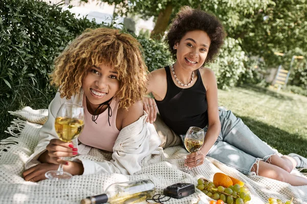 Namoradas americanas africanas com copos de vinho sorrindo para a câmera perto de frutas frescas, piquenique, verão — Fotografia de Stock