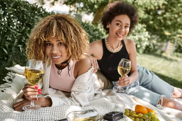 Mujeres afroamericanas felices con copas de vino mirando a la cámara, picnic de verano en el parque - foto de stock