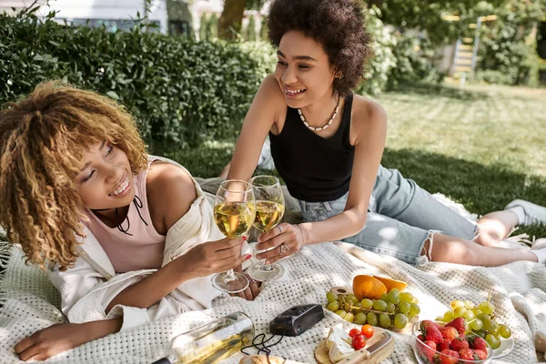 Alegres novias afroamericanas tintineando copas de vino cerca de frutas frescas, picnic de verano en el parque - foto de stock