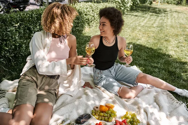 Fröhliche afrikanisch-amerikanische Frau mit Weingläsern in der Nähe von Früchten und Freundin, Picknick im Park — Stockfoto
