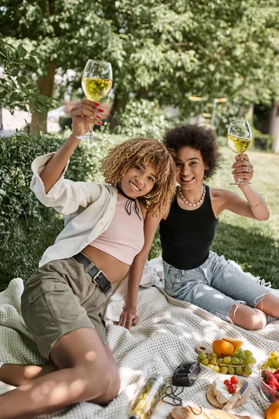 Gioiosa donna afroamericana brindare con bicchiere di vino vicino fidanzata durante il picnic estivo nel parco — Foto stock