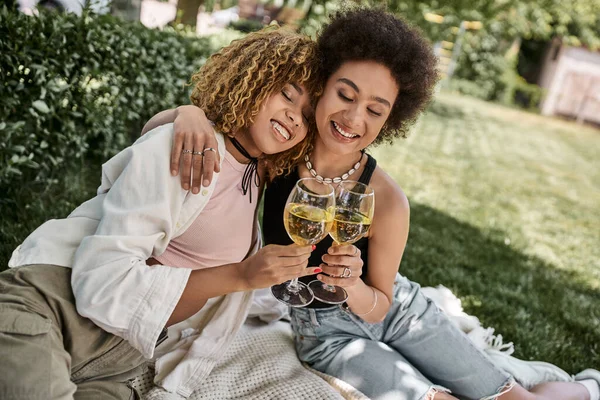 Amigas afroamericanas positivas con los ojos cerrados tintineo copas de vino, picnic de verano en el parque - foto de stock