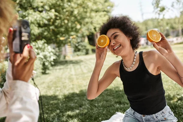 Щаслива афроамериканська жінка позує з половиною апельсина біля дівчини з старовинною камерою в парку — стокове фото