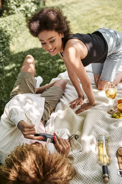 Positive african american woman posing near girlfriend with vintage camera on blanket in park — Stock Photo