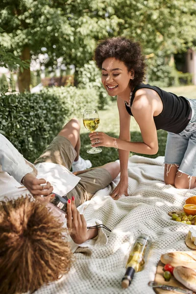 Mulher americana africana feliz com copo de vinho perto da namorada com câmera vintage no cobertor no parque — Fotografia de Stock
