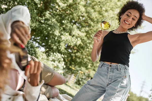 Vista ritagliata di donna afro-americana con bicchiere di vino vicino fidanzata con fotocamera vintage nel parco — Foto stock