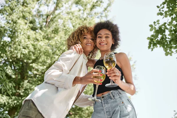 Novias afroamericanas despreocupadas con copas de vino abrazándose en el parque, picnic de verano - foto de stock