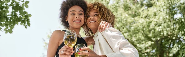 Picnic de verano, alegres novias afroamericanas con copas de vino abrazándose en el parque, pancarta - foto de stock