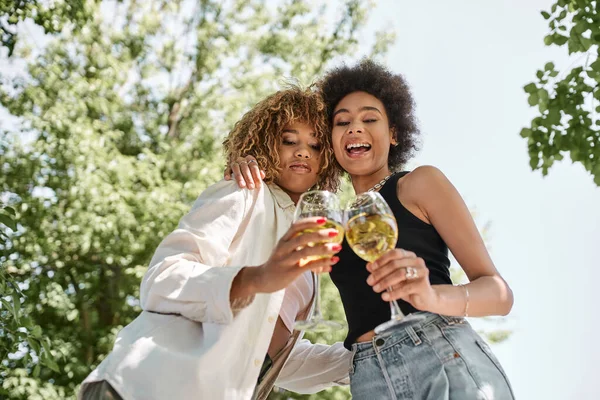 Riéndose novias afroamericanas abrazando y tintineando copas de vino, parque, verano, picnic - foto de stock