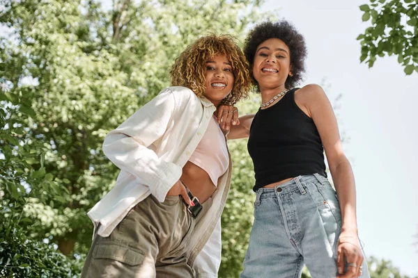 Overjoyed and stylish african american girlfriends looking at camera in summer park, low angle view — Stock Photo