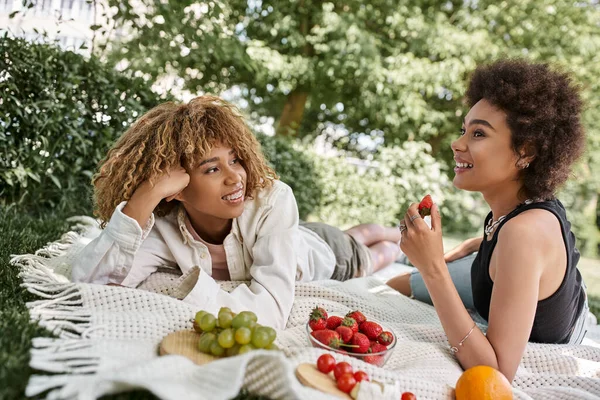 Счастливые африканские американские девушки разговаривают рядом со свежими фруктами на одеяле, летний пикник в парке — стоковое фото