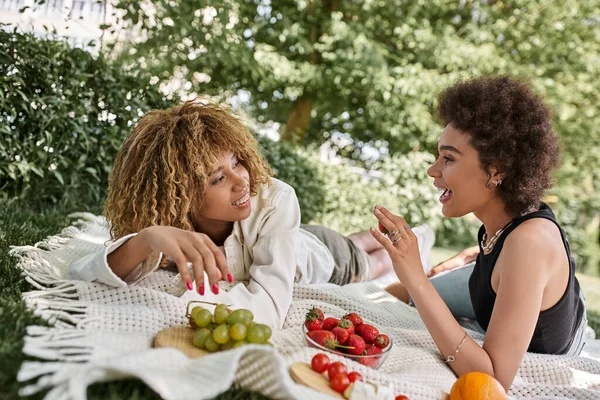 Picnic de verano, mujer afroamericana emocionada hablando con su novia cerca de frutas frescas en manta - foto de stock