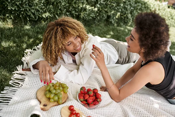 African american woman holding fresh strawberry near girlfriend during picnic in park — Stock Photo