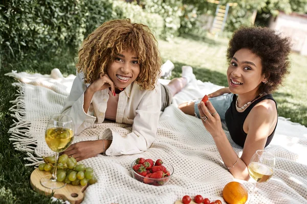 Picnic de verano, novias afroamericanas positivas mirando a la cámara cerca de frutas y copa de vino - foto de stock