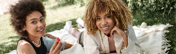 Donna afroamericana con fragola fresca vicino fidanzata sorridente alla macchina fotografica, parco estivo, banner — Foto stock