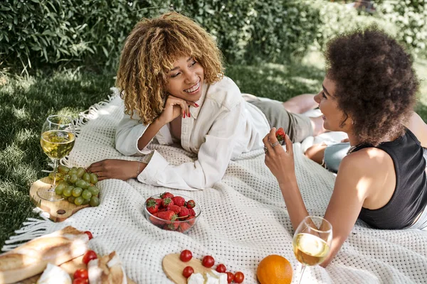 Picnic de verano, novias afroamericanas complacidas hablando cerca de frutas y copas de vino en el parque - foto de stock