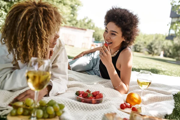 Unbekümmerte Afroamerikanerin hält Erdbeere in der Hand und spricht mit Freundin beim Sommerpicknick — Stockfoto