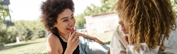 Sonriente mujer afroamericana con fresa hablando con su novia en el parque, picnic, pancarta - foto de stock