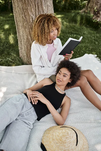 Усміхнена афроамериканська жінка читає книгу біля дівчини відпочиває на ковдрі в літньому парку — стокове фото