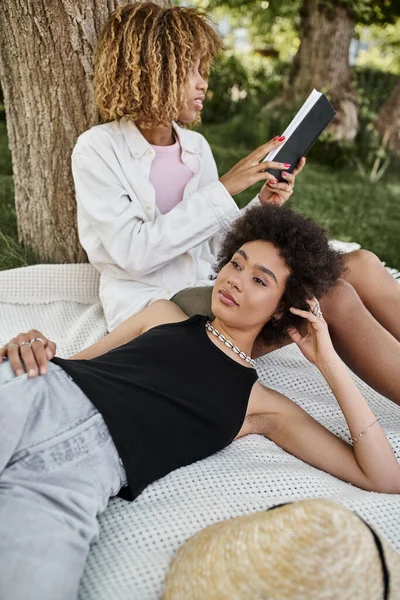 Молодая африканская американка лежит рядом с девушкой, читает книгу о одеяле, летний отдых в парке — стоковое фото