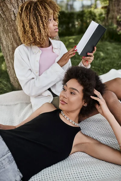 Ocio en el parque de verano, mujer afroamericana leyendo libro cerca de novia puesta en manta - foto de stock