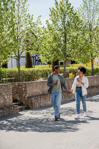 Smiling african american couple holding hands and coffee to go while walking on urban street — Stock Photo