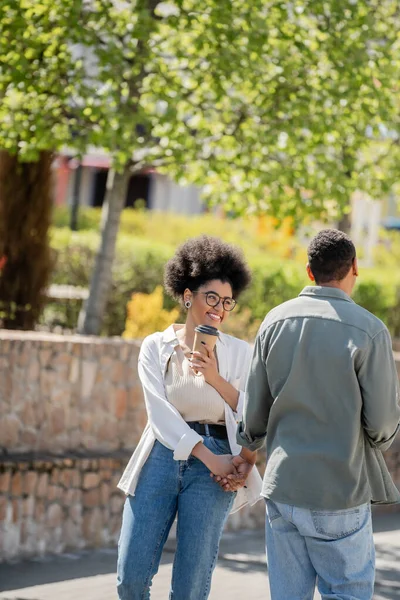 Cheerful african american woman holding coffee to go and hand of boyfriend on urban street — Stock Photo