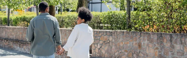 Веселая африканская американка, держащая за руку бойфренда во время прогулки по городской улице, баннер — стоковое фото